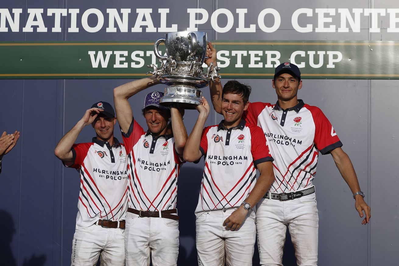 A group of gentlemen in white outfits with red sleeves lift a trophy over their heads in front of a background that reads National Polo Center