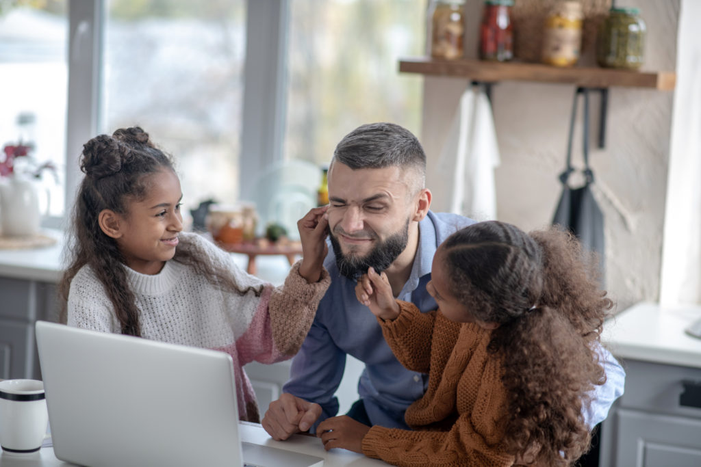 Cheerful curly-haired daughters having fun with daddy while he's trying to work on his laptop.