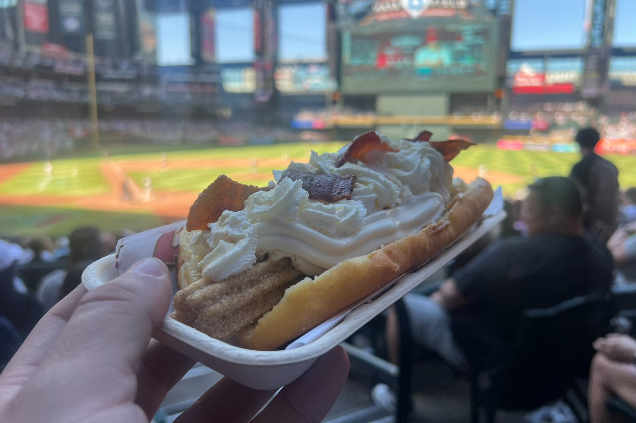 My hand in the bottom left corner, holding a churro dog at an angle with the Chase Field jumbotron and field out of focus in the background